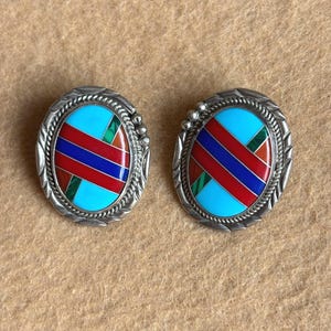 May include: A pair of oval-shaped stud earrings. Each earring features a turquoise center with red and blue stripes, accented with green. The earrings are set in a silver-toned metal with a decorative border.