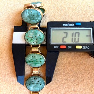 May include: A gold-toned bracelet with oval, carved jade stones. The bracelet is measured with a digital caliper, showing a width of 21.0 mm. The jade stones have a light green color and are connected by gold links.