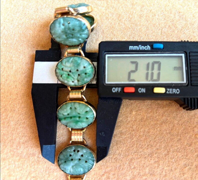 May include: A gold-toned bracelet with oval, carved jade stones. The bracelet is measured with a digital caliper, showing a width of 21.0 mm. The jade stones have a light green color and are connected by gold links.