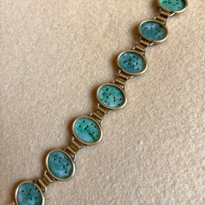 May include: A gold-toned bracelet featuring six oval jade stones with dark green speckles. The jade stones are set in gold-colored frames and linked together with gold-toned metal. The bracelet is displayed on a beige surface.