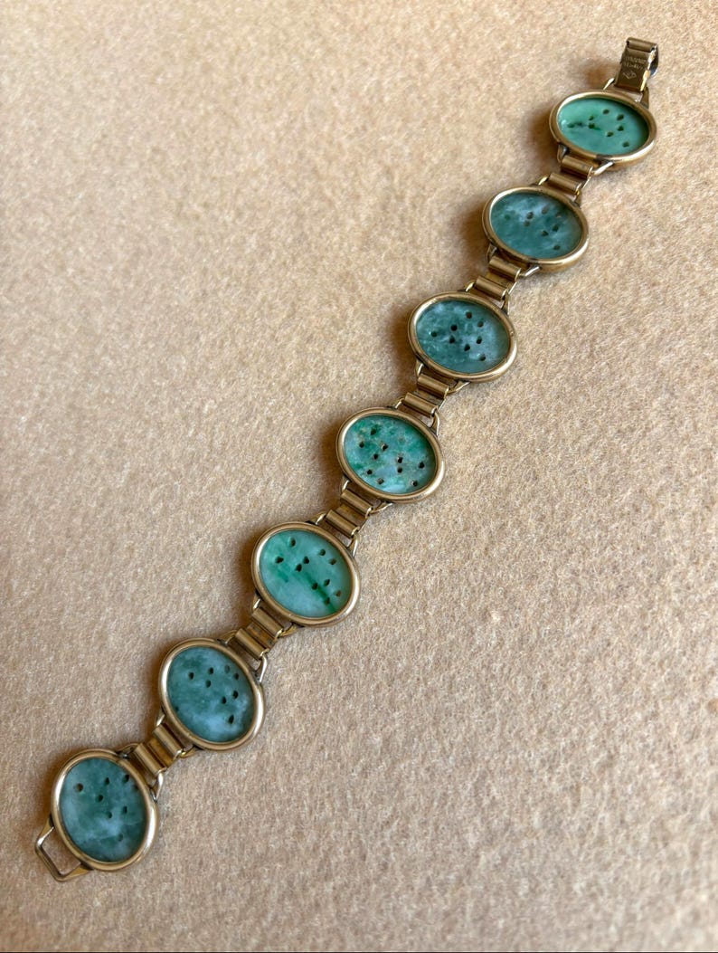May include: A gold-toned bracelet featuring six oval jade stones with dark green speckles. The jade stones are set in gold-colored frames and linked together with gold-toned metal. The bracelet is displayed on a beige surface.