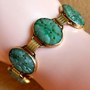 May include: A gold-toned bracelet featuring oval jade stones with carved floral designs. The jade is a vibrant green color, and the bracelet is connected with gold-colored links. The bracelet is displayed on a light pink surface.
