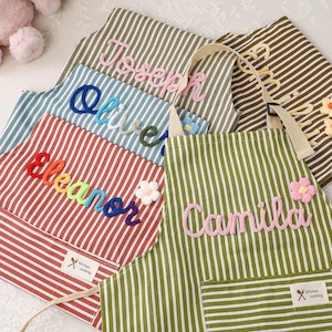 May include: Striped aprons in various colors, each embroidered with a name in a cursive font. The aprons have a pocket and a tag that says "kitchen cooking". Some aprons have floral embellishments.