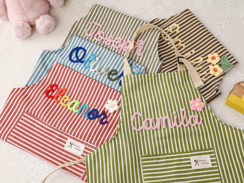 May include: Striped aprons in various colors, each embroidered with a name in a cursive font. The aprons have a pocket and a tag that says "kitchen cooking". Some aprons have floral embellishments.