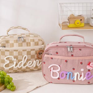 Op de afbeelding: Twee corduroy lunchtassen. Eén is beige en geruit met de naam "Eiden" in witte letters. De andere is roze met een aardbeienpatroon en de naam "Bonnie" in kleurrijke letters. Beide hebben handvatten en ritsen.