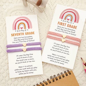 Może przedstawiać: Dwie bransoletki przyjaźni na kartach, jedna fioletowa i jedna różowa, ze złotymi zawieszkami w kształcie serca. Na kartach widnieją napisy "First Day of Seventh Grade" i "First Grade" z tęczowymi wzorami. Ołówki i notatnik w tle.