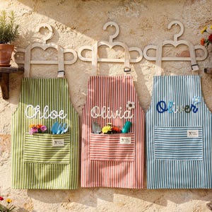 May include: Three striped aprons, green, red, and blue, each with a name embroidered on them: Chloe, Olivia, and Oliver. Each apron has a pocket filled with gardening tools and flowers. They hang on wooden hangers.