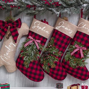 Peut inclure: Chaussettes de Noël à carreaux rouges et noirs, avec des hauts en toile de jute. Une chaussette en forme d'os porte le nom "Markey". Les trois autres sont personnalisées avec "Sophia", "James" et "Christina". Décorées de verdure et de nœuds.