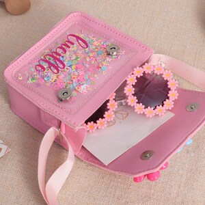 May include: A pink crossbody bag with a clear front pocket filled with colorful confetti and the name Lucille in script. The bag features a magnetic snap closure and contains a pair of round sunglasses with a pink daisy flower frame border.