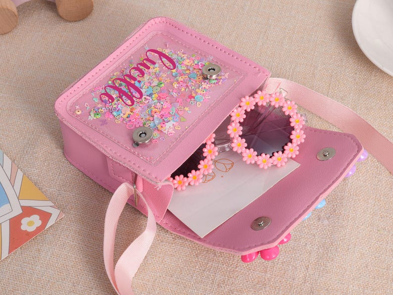 May include: A pink crossbody bag with a clear front pocket filled with colorful confetti and the name Lucille in script. The bag features a magnetic snap closure and contains a pair of round sunglasses with a pink daisy flower frame border.