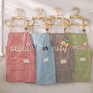 May include: Four striped aprons in red, blue, brown, and green hang on wooden hangers. Each apron has a pocket and a name embroidered on it: Olivia, River, Emily, and Camila. Decorative floral accents are attached to some of the names.