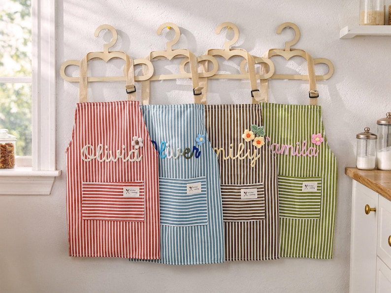 May include: Four striped aprons in red, blue, brown, and green hang on wooden hangers. Each apron has a pocket and a name embroidered on it: Olivia, River, Emily, and Camila. Decorative floral accents are attached to some of the names.