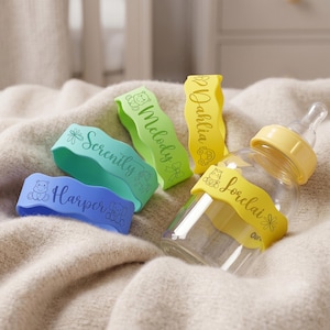 Pode incluir: Uma mamadeira com tampa amarela e uma faixa amarela com o nome "Lorclai" impresso. Há também três faixas coloridas com os nomes "Harper", "Serenity" e "Melody" impressos. As faixas são azuis, verdes e amarelas.