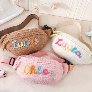 May include: Three plush fanny packs in brown, white, and pink. Each pack has a different name embroidered in colourful thread: "Lucas", "Layla", and "Chloe". The bags have adjustable straps and black buckles.
