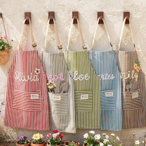 May include: Five striped aprons hang on a wall, each with a different color scheme: red, gray, green, blue, and brown. Each apron has a pocket and a name embroidered on it. A hanging flower pot is also visible.