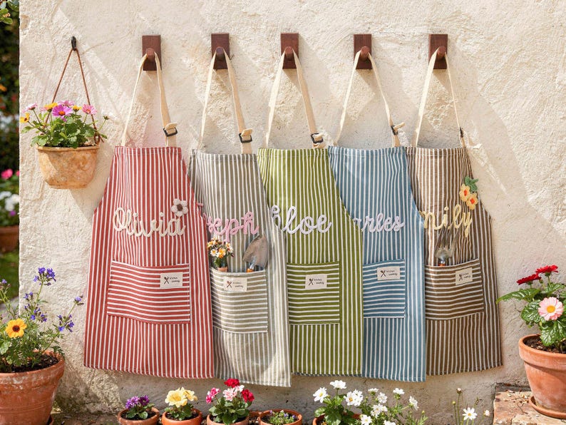 May include: Five striped aprons hang on a wall, each with a different color scheme: red, gray, green, blue, and brown. Each apron has a pocket and a name embroidered on it. A hanging flower pot is also visible.