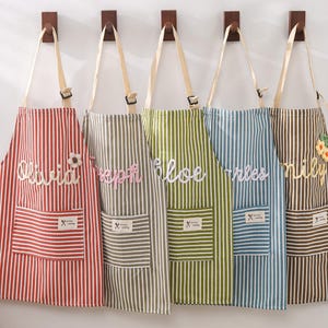 May include: Five striped aprons in red, gray, green, blue, and brown hang on wooden hooks. Each apron has a pocket and a name embroidered on it: Olivia, Joseph, Chloe, Charles, and Lily. The aprons have adjustable straps.