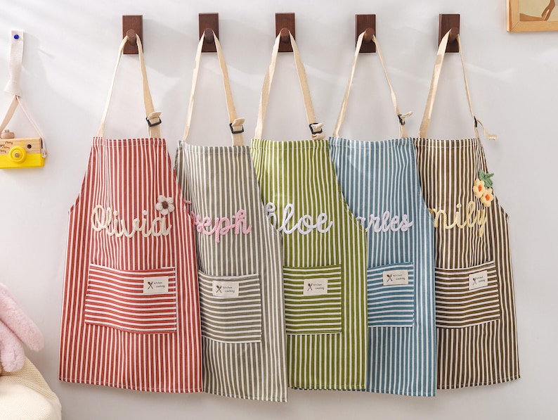 May include: Five striped aprons in red, gray, green, blue, and brown hang on wooden hooks. Each apron has a pocket and a name embroidered on it: Olivia, Joseph, Chloe, Charles, and Lily. The aprons have adjustable straps.