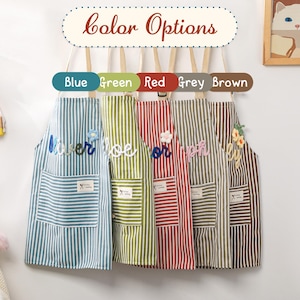 May include: Five striped aprons in blue, green, red, grey, and brown hang on a wooden rack. Each apron has a pocket and a decorative element. The sign above reads "color Options".