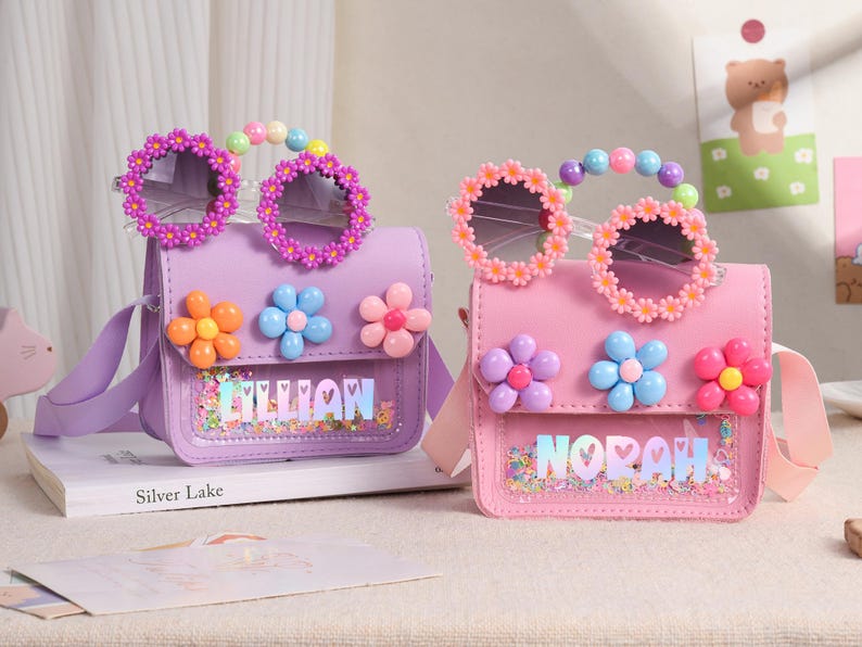 May include: Two small rectangular handbags in lavender and pink, each featuring a clear front pocket filled with colorful confetti. The bags are decorated with three-dimensional flower appliques and have a handle made of pastel beads.