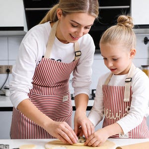 May include: Two aprons with red and white stripes are worn by two people in a kitchen. The aprons have beige straps and are being used while cutting dough with cookie cutters. The aprons are part of a baking set.