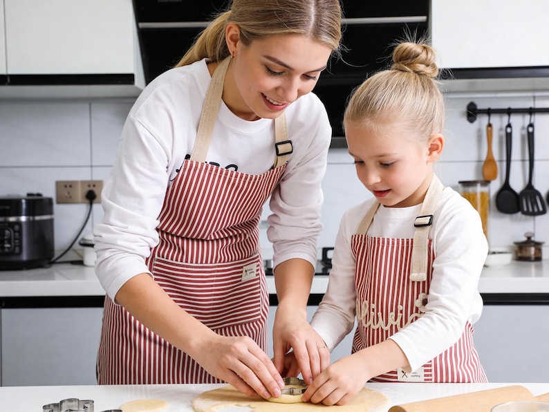 May include: Two aprons with red and white stripes are worn by two people in a kitchen. The aprons have beige straps and are being used while cutting dough with cookie cutters. The aprons are part of a baking set.