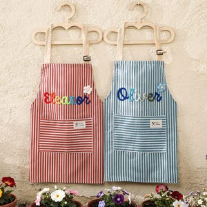 May include: Two striped aprons, one red and one blue, hang on wooden hangers. The red apron has the name "Eleanor" embroidered in colorful letters, and the blue apron says "Oliver". Both aprons have a pocket and a floral embellishment.