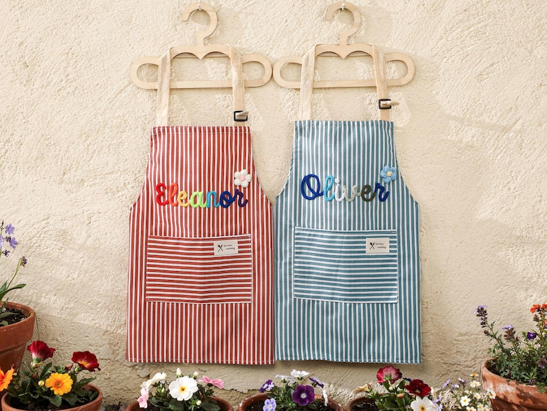 May include: Two striped aprons, one red and one blue, hang on wooden hangers. The red apron has the name "Eleanor" embroidered in colorful letters, and the blue apron says "Oliver". Both aprons have a pocket and a floral embellishment.