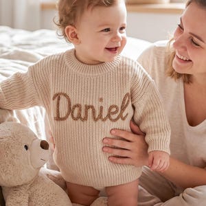 以下が含まれることがあります： 胸元に茶色の筆記体でDanielという名前が刺繍された、ベージュの長袖ニットセーター。このセーターは、リブ編みのクルーネック、リブ編みの袖口、リブ編みの裾が特徴で、厚みのある縦方向のニットパターンで構成されています。