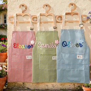 May include: Three personalized aprons with names embroidered on them. The aprons are red, green, and blue with white stripes. Each apron has a pocket and is hanging on a wooden hanger. The names are Eleanor, Camila, and Oliver.