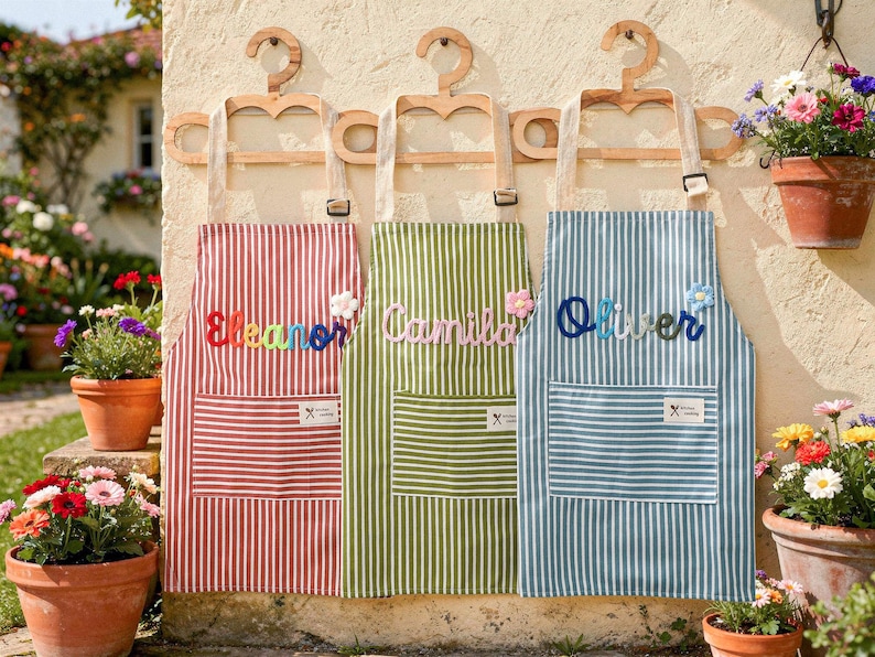 May include: Three personalized aprons with names embroidered on them. The aprons are red, green, and blue with white stripes. Each apron has a pocket and is hanging on a wooden hanger. The names are Eleanor, Camila, and Oliver.