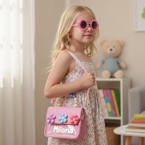 May include: A pink handbag featuring a clear front pocket with the name Milana printed in white, adorned with three colorful flower embellishments and a handle made of multi-colored beads, accompanied by a matching pink shoulder strap.