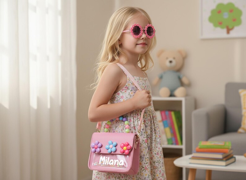 May include: A pink handbag featuring a clear front pocket with the name Milana printed in white, adorned with three colorful flower embellishments and a handle made of multi-colored beads, accompanied by a matching pink shoulder strap.