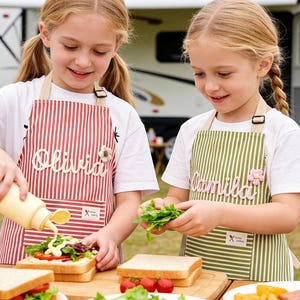 May include: Two young girls wearing aprons with names on them prepare sandwiches outdoors. One apron is red and white striped, the other is green and white striped. The aprons have a flower and the text "Olivia" and "Camila".