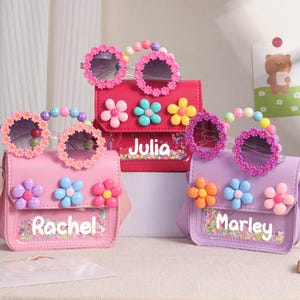 May include: Three small rectangular crossbody bags in pink, red, and purple, each featuring a clear front pocket with colorful sequins, a name printed in white, three large plastic flower embellishments, and matching daisy-rimmed sunglasses.