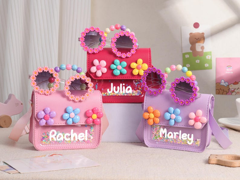 May include: Three small rectangular crossbody bags in pink, red, and purple, each featuring a clear front pocket with colorful sequins, a name printed in white, three large plastic flower embellishments, and matching daisy-rimmed sunglasses.