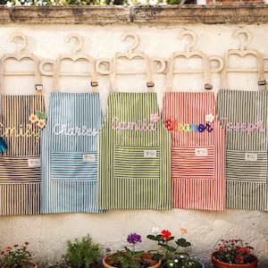 May include: Five striped aprons in brown, blue, green, red, and gray, each personalized with a name in cursive. The aprons hang on wooden hangers against a white wall, with potted plants in the foreground. Each apron has a front pocket.