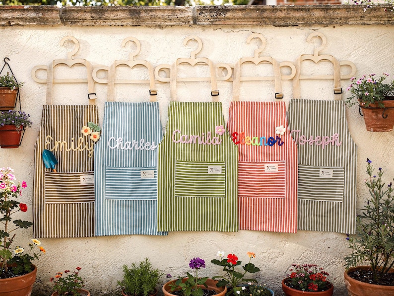 May include: Five striped aprons in brown, blue, green, red, and gray, each personalized with a name in cursive. The aprons hang on wooden hangers against a white wall, with potted plants in the foreground. Each apron has a front pocket.