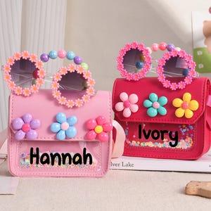 May include: Two small rectangular crossbody bags in light pink and dark pink, each featuring a clear front pocket with colorful confetti, the names Hannah and Ivory, three dimensional flower accents, and matching flower-rimmed sunglasses.