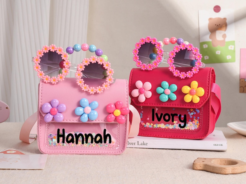 May include: Two small rectangular crossbody bags in light pink and dark pink, each featuring a clear front pocket with colorful confetti, the names Hannah and Ivory, three dimensional flower accents, and matching flower-rimmed sunglasses.