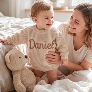 Pode incluir: Uma camisola de malha bege de manga comprida com o nome Daniel bordado em letra cursiva castanha no peito. A camisola apresenta um decote redondo canelado, punhos canelados e uma bainha canelada, proporcionando um aspeto texturado.