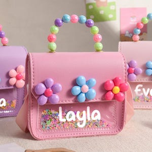 May include: A pink handbag featuring a clear front pocket filled with colorful confetti, a handle made of multi-colored beads, and three plastic flower embellishments. The name Layla is printed in white on the clear section of the bag.