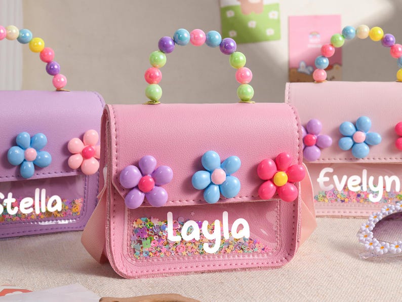 May include: A pink handbag featuring a clear front pocket filled with colorful confetti, a handle made of multi-colored beads, and three plastic flower embellishments. The name Layla is printed in white on the clear section of the bag.