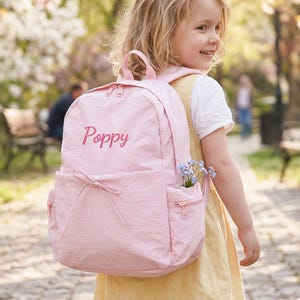 Könnte beinhalten: Ein hellrosa Rucksack mit dem Namen "Poppy" auf der Vorderseite gestickt. Der Rucksack hat eine Vordertasche mit Schleife und eine Seitentasche mit blauen Blumen. Das Kind trägt ein gelbes Kleid.