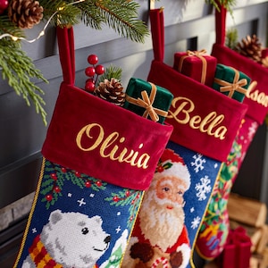 Könnte beinhalten: Drei Weihnachtsstrümpfe mit roten Samtstulpen und den gold gestickten Namen Olivia, Bella sowie einem Teilnamen. Die Strümpfe zeigen Stickereimotive eines Eisbären, des Weihnachtsmanns und festliche Muster, verziert mit kleinen Geschenkboxen.