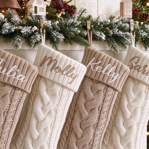 Op de afbeelding: Een rij van vier gebreide kerstsokken hangend aan een schoorsteenmantel. De sokken hebben een kabelpatroon in afwisselende crème en beige kleuren. Elke omslag is geborduurd met de namen Julia, Molly, Kelly en Sara.