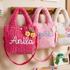 Peut inclure: Trois sacs fourre-tout en velours côtelé avec bandoulières, chacun arborant un prénom brodé en écriture cursive et une petite applique florale. Les sacs sont de couleur rose vif, rose clair et blanc, avec des fermetures à pression blanches.
