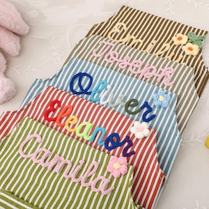 May include: A stack of striped aprons in various colors, each embroidered with a name in a different color. The names include Camila, Eleanor, Oliver, Joseph, and Emily. Decorative floral accents are also present.