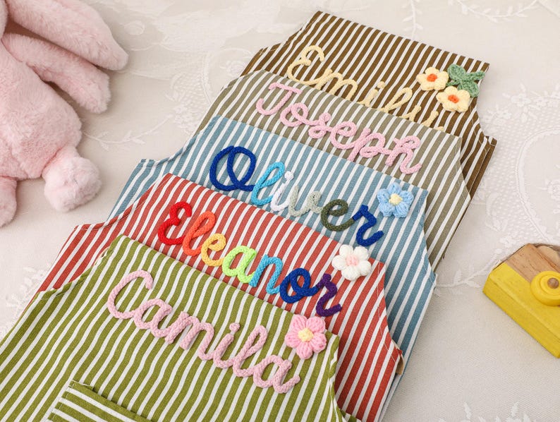 May include: A stack of striped aprons in various colors, each embroidered with a name in a different color. The names include Camila, Eleanor, Oliver, Joseph, and Emily. Decorative floral accents are also present.