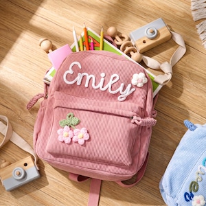 May include: A pink corduroy backpack featuring the name Emily embroidered in white script, a small white crocheted flower, and a front pocket decorated with two pink crocheted flowers with green leaves, containing coloured pencils and a book.